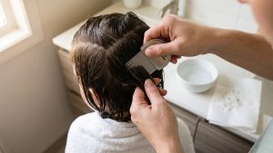 Parent using a fine-toothed stainless-steel lice comb on a child's wet, sectioned hair in a home bathroom.
