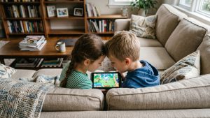 Two children sitting side-by-side on a couch with their heads close together while sharing a tablet, illustrating that head lice spread by direct head-to-head contact rather than by jumping or flying.