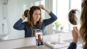 Adult woman in a sunlit bathroom checking her own hair for head lice with a fixed wall mirror, a hand mirror behind her head, and a smartphone in a stand showing the selfie-camera view of the back of her scalp on the counter beside a fine-toothed metal lice comb.