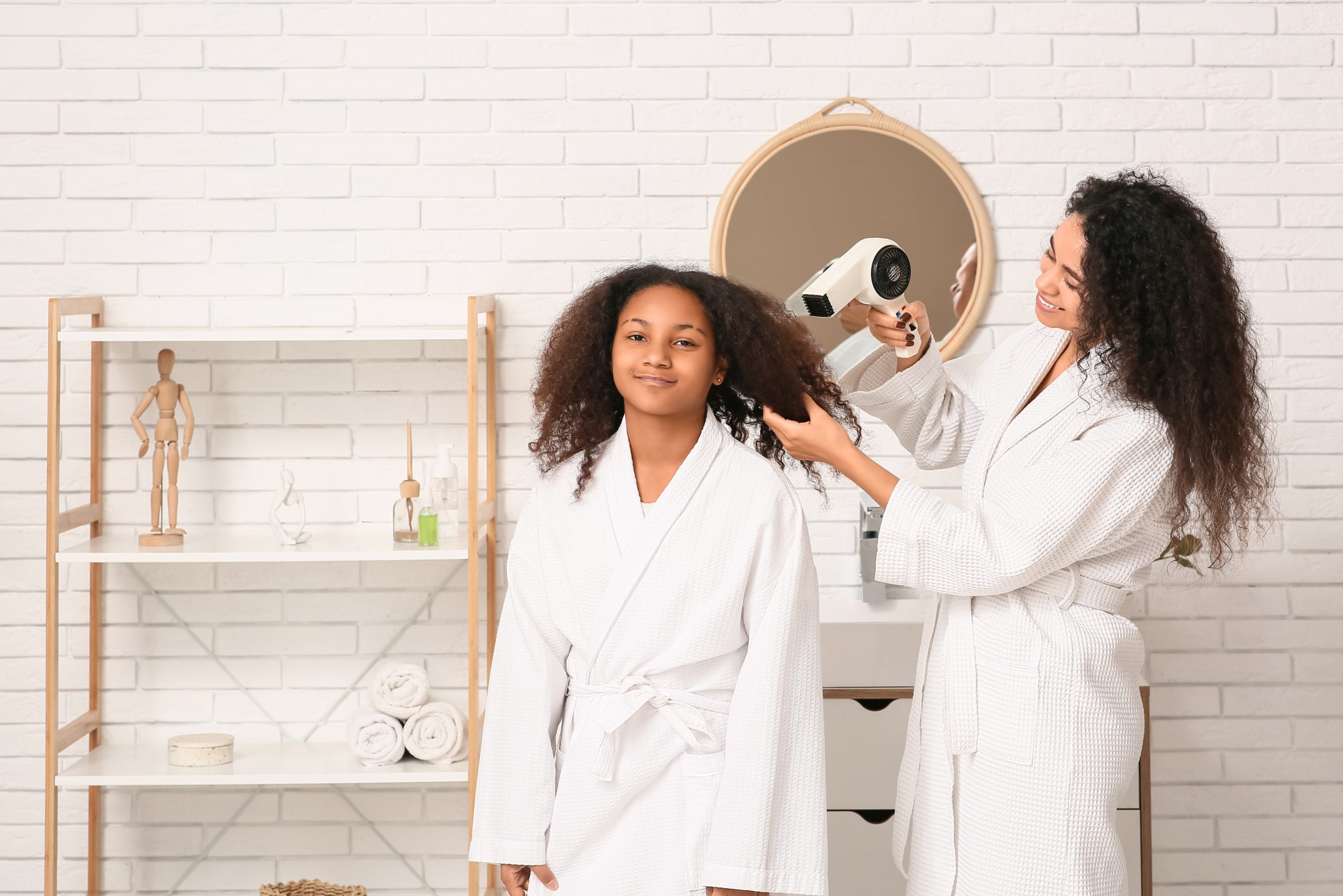 Mother carefully blow-drying a school-age child's curly hair in a sunlit bathroom with a fine-tooth nit comb and conditioner bottle on the counter.