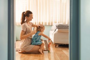 Mother gently brushing her young daughter's hair at home on a couch while thinking about how long head lice can survive on pillows, bedding, and household surfaces