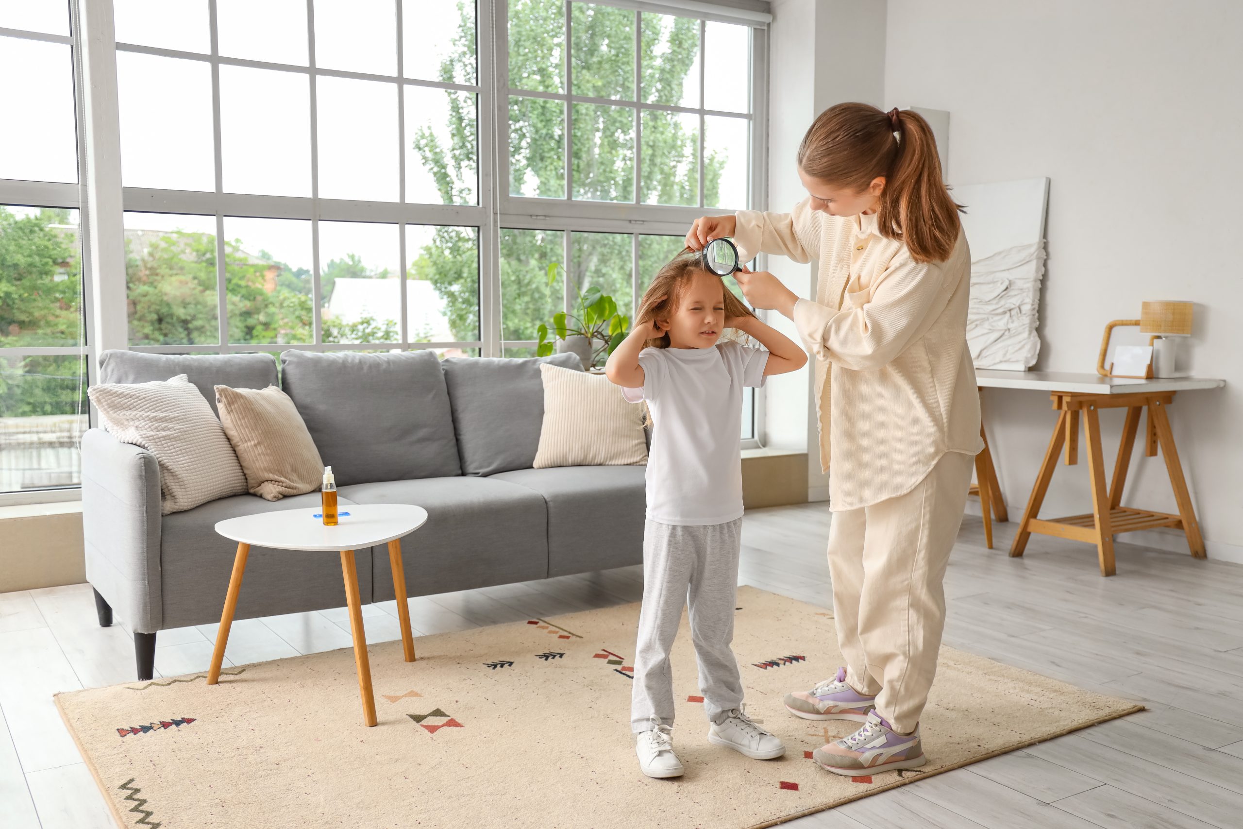 A parent checking a young child's scalp with a magnifier during an at-home head lice check in a bright living room.