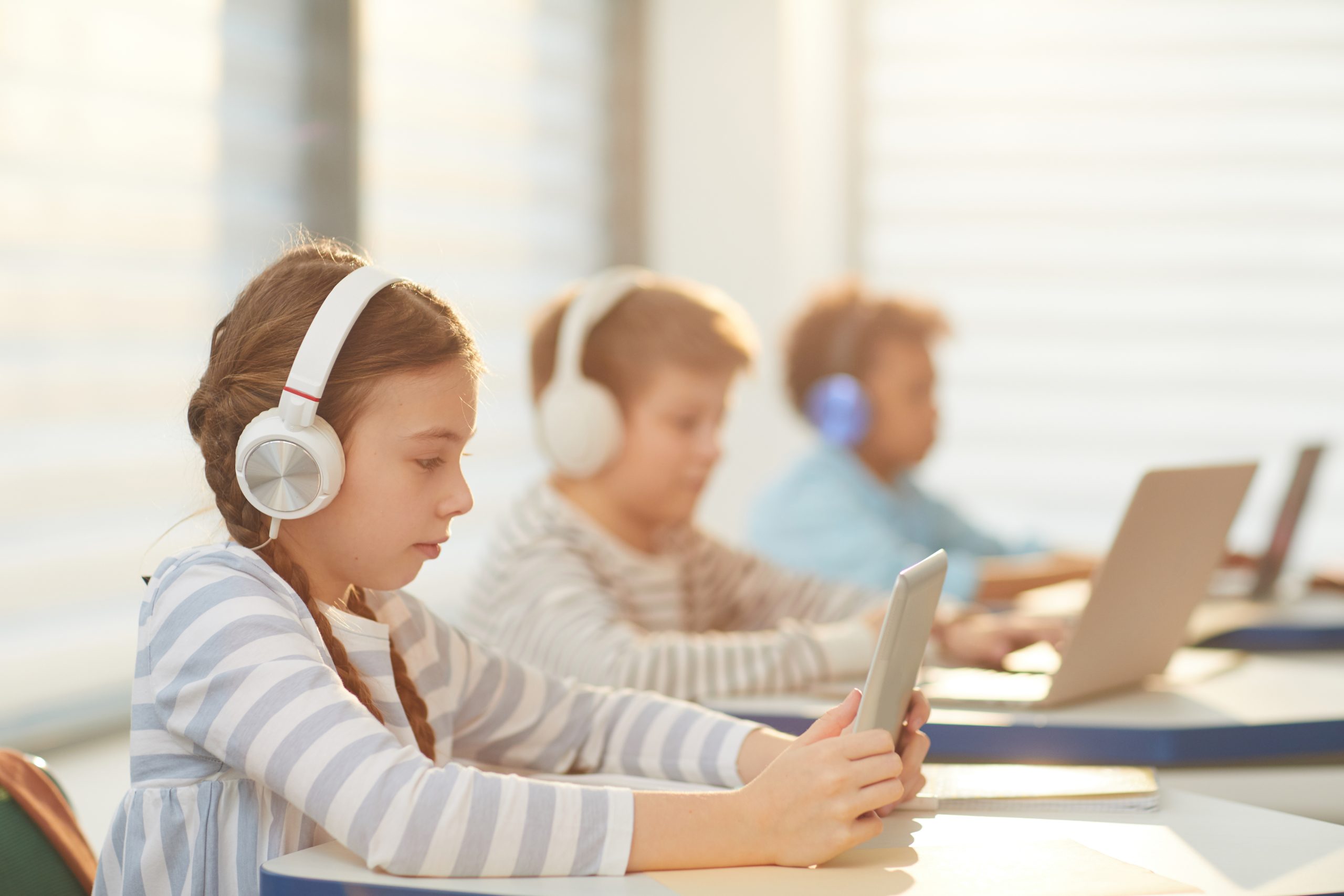 Three school-age children at classroom desks wearing over-ear headphones while working on tablets and laptops