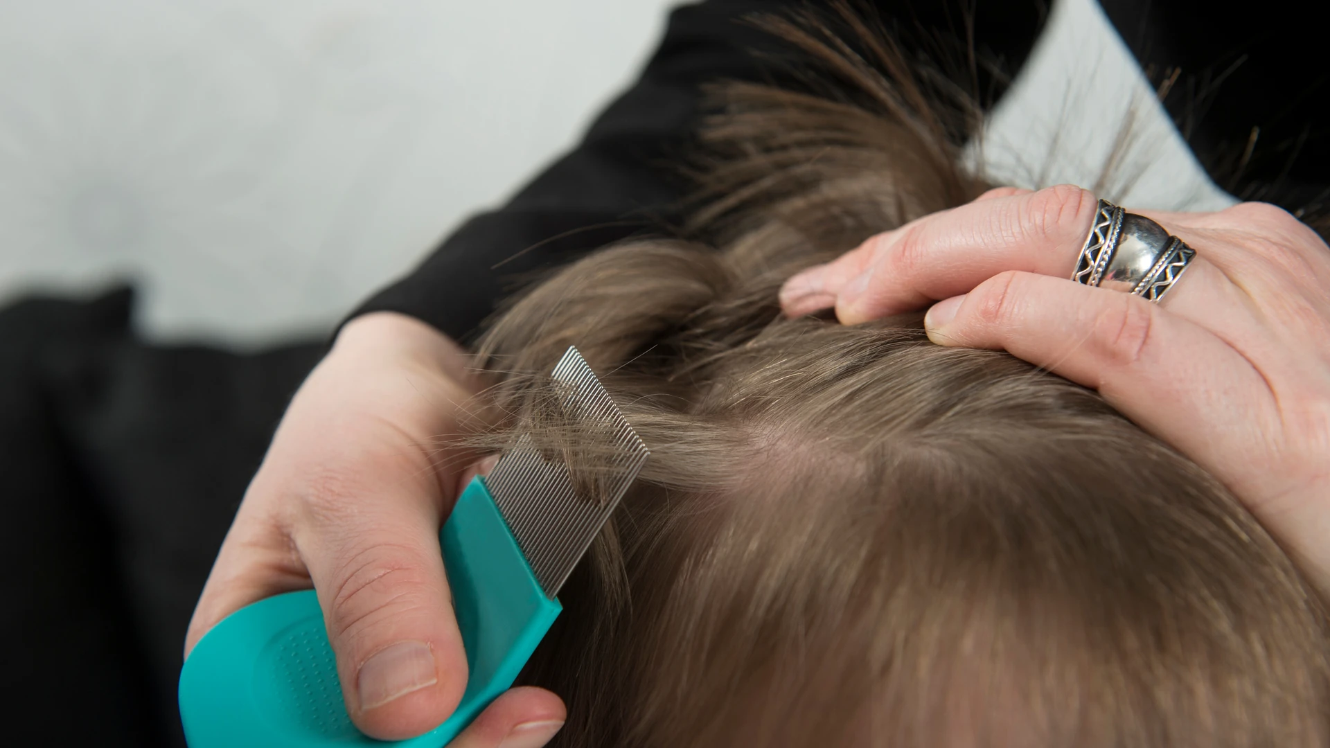 Hands holding light brown hair aside while using a green-handled fine-tooth comb.