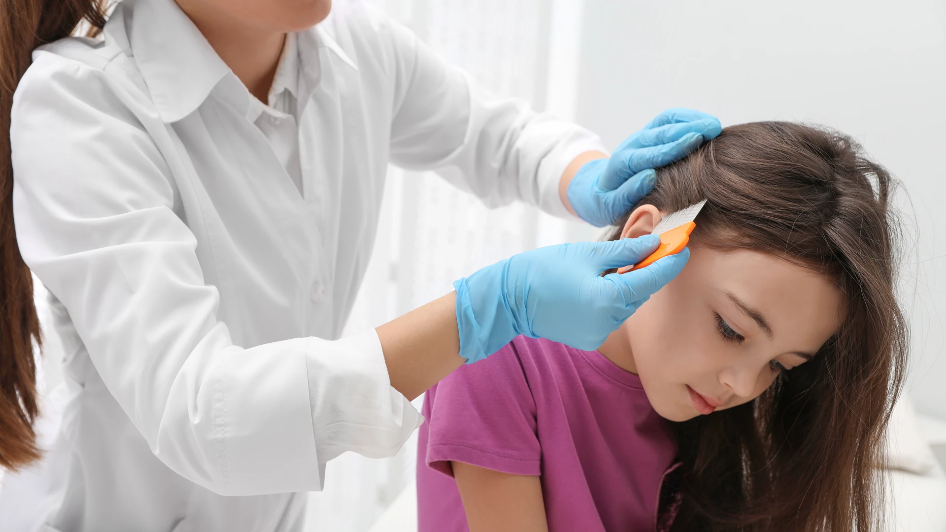 Blue-gloved hands using an orange fine-tooth comb near an ear while holding the hair aside.